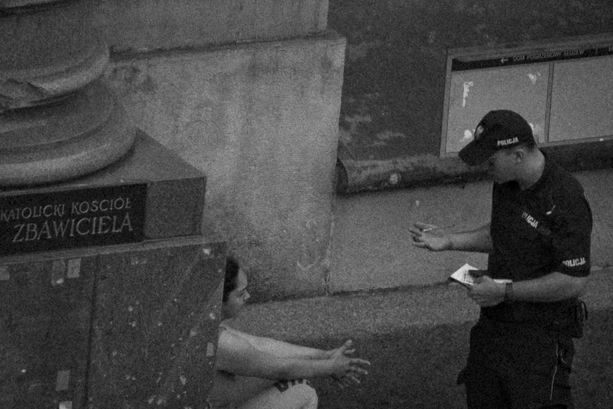 Police officer holding paperwork while a seated woman reaches toward him from behind a stone column